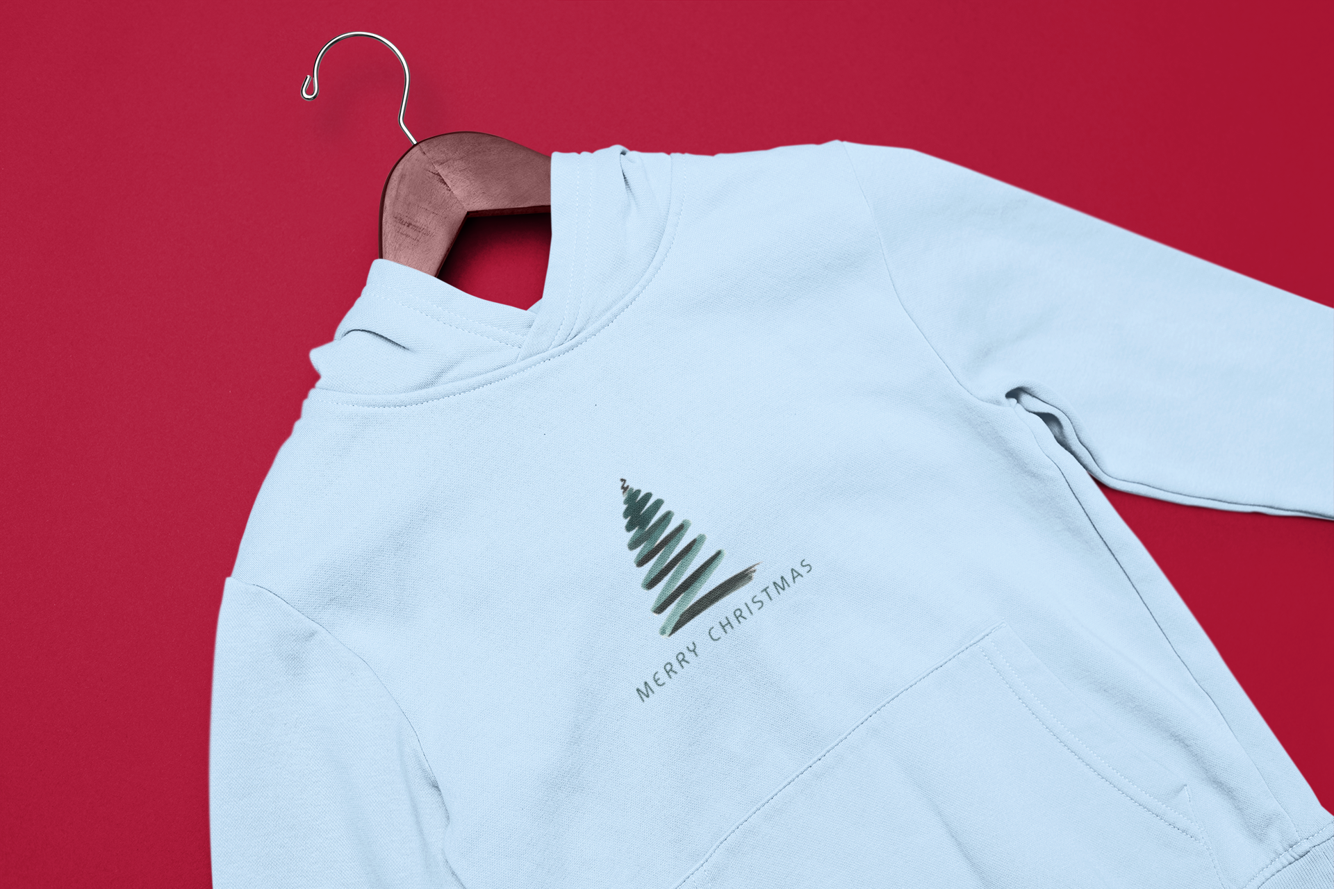 A white hoodie with a Christmas tree design and the text "MERRY CHRISTMAS" printed on it, hanging on a wooden hanger against a red background.