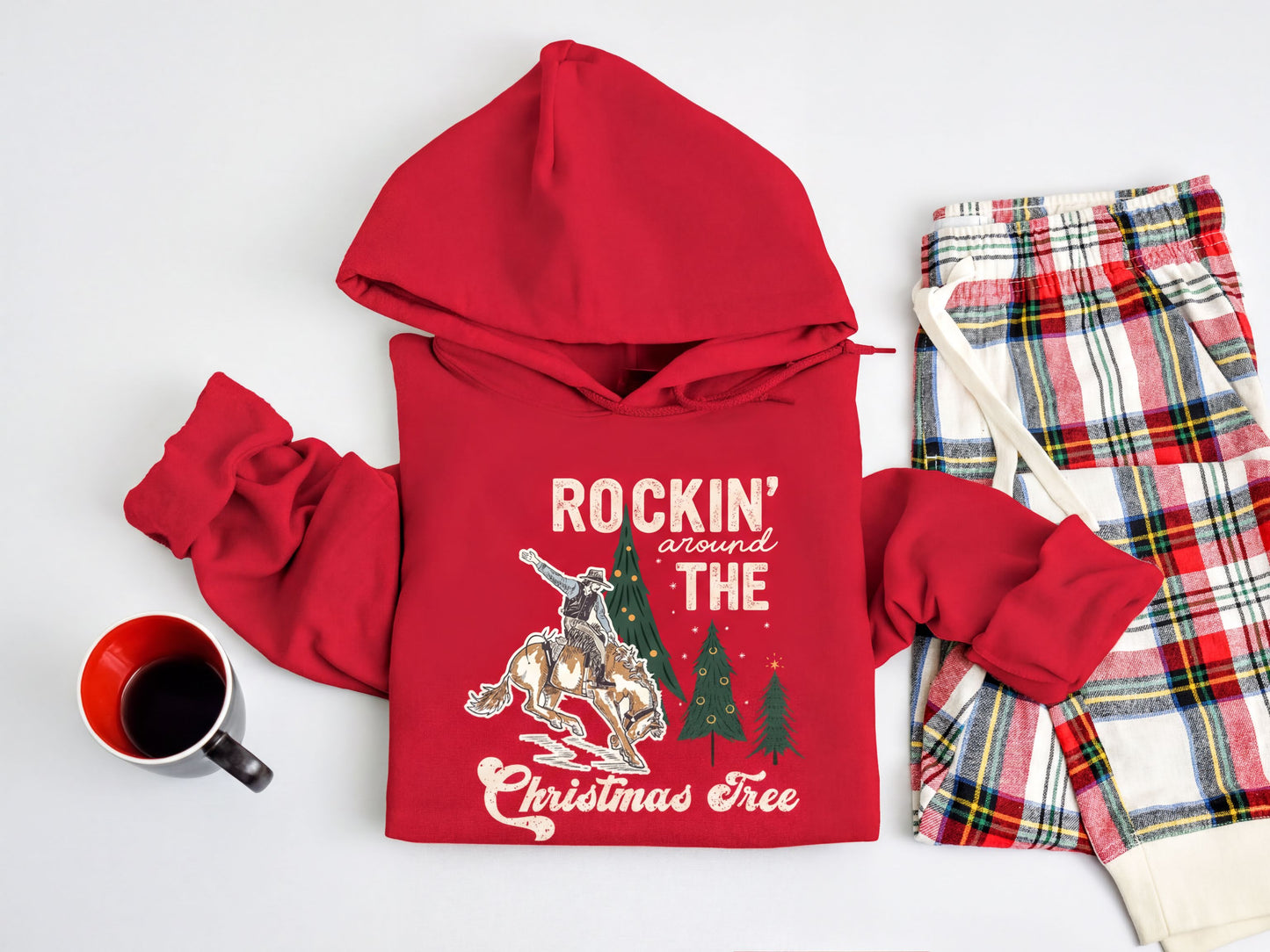 A red hoodie with a Christmas tree design and the text "ROCKIN' around THE CHRISTMAS Tree" is displayed on a white background, along with a pair of plaid pajama pants and a mug.