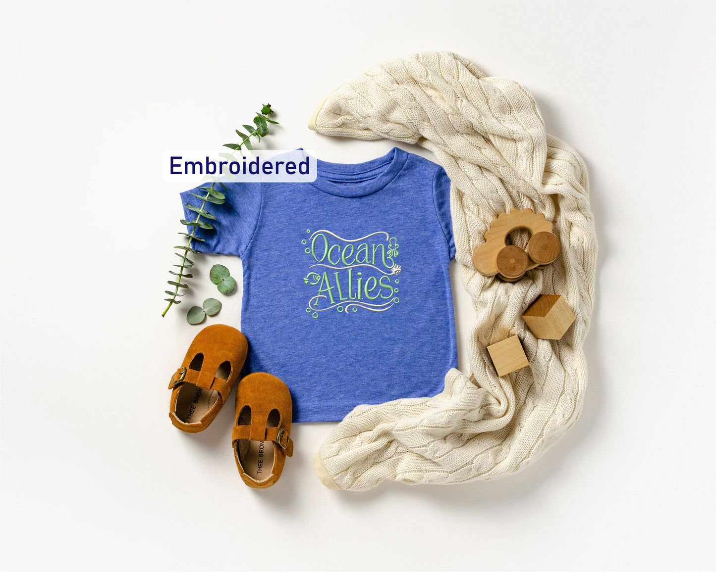 a blue t-shirt with the text "Ocean Allies" embroidered on it, along with a pair of brown sandals, a white blanket, and some wooden toys.
