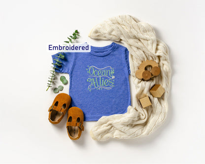 a blue t-shirt with the text "Ocean Allies" embroidered on it, along with a pair of brown sandals, a white blanket, and some wooden toys.