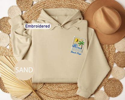 a beige hoodie with a beach-themed patch, a brown hat, and a fan, all arranged on a woven mat.