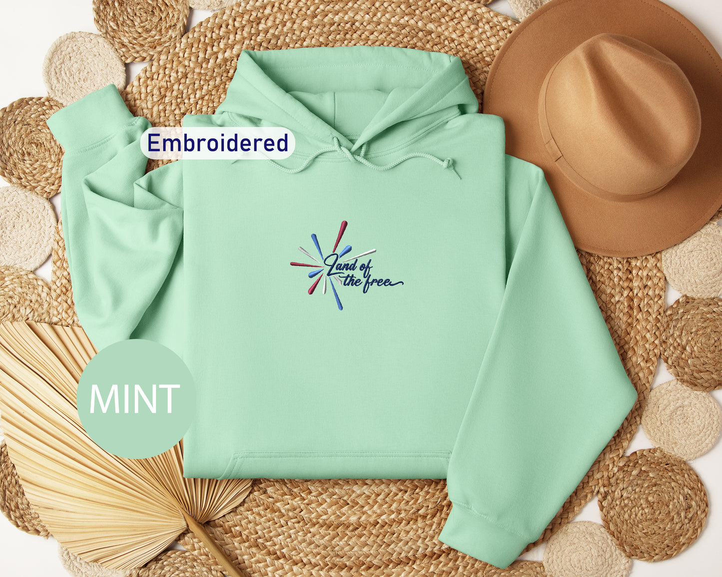a mint green hoodie with a colorful embroidered design, a brown hat, and a straw hat, all arranged on a woven mat.