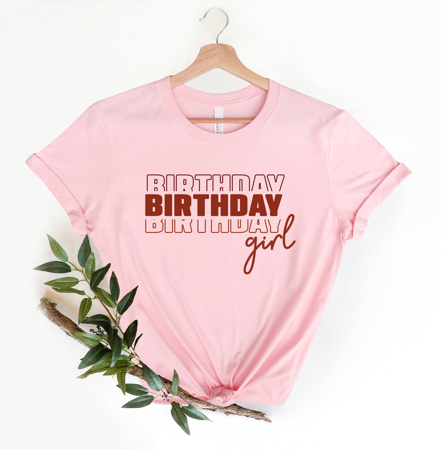 A pink t-shirt with the text "BIRTHDAY BIRTHDAY girl" printed on it, along with a wooden hanger.