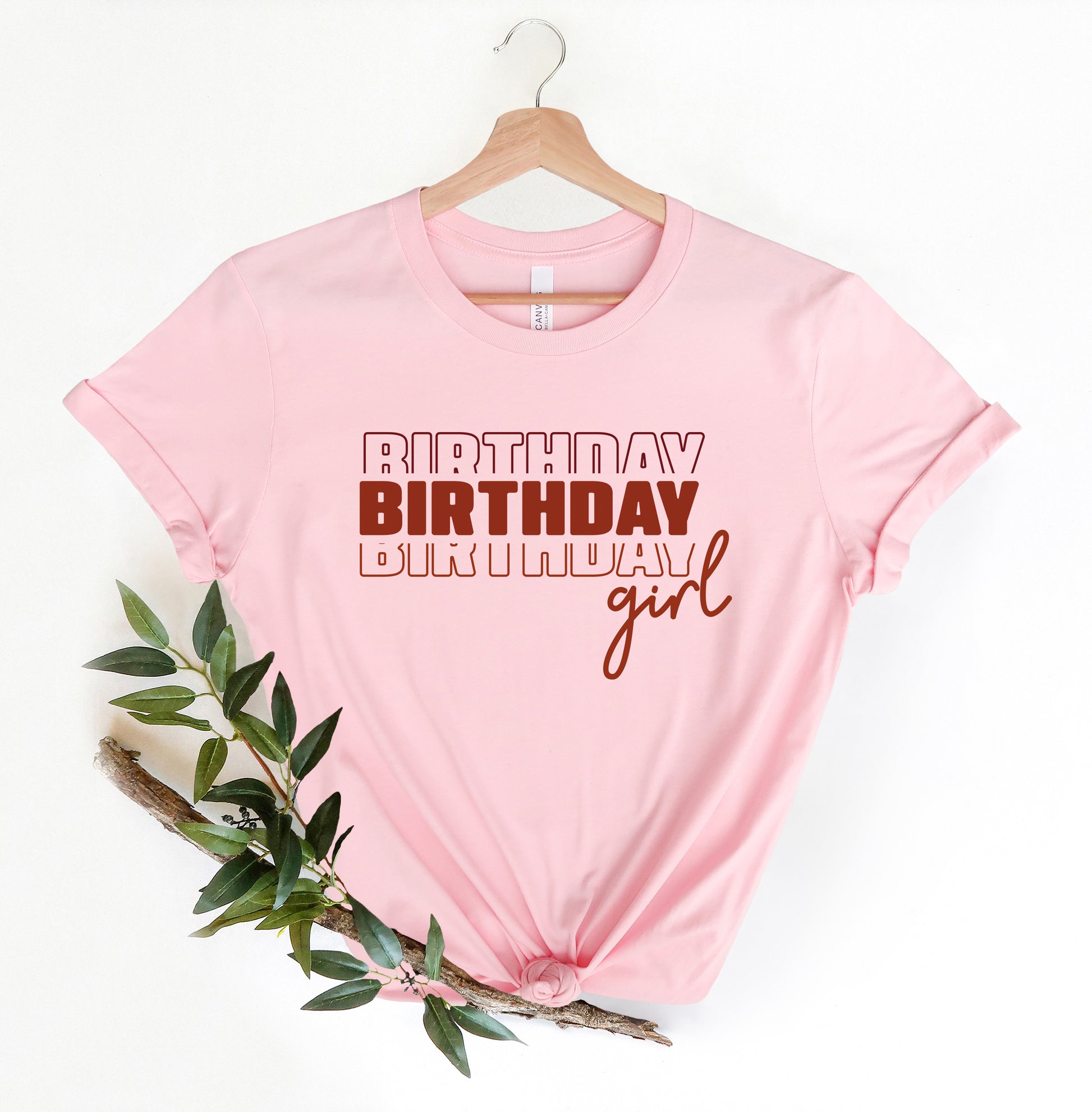 A pink t-shirt with the text "BIRTHDAY BIRTHDAY girl" printed on it, along with a wooden hanger.
