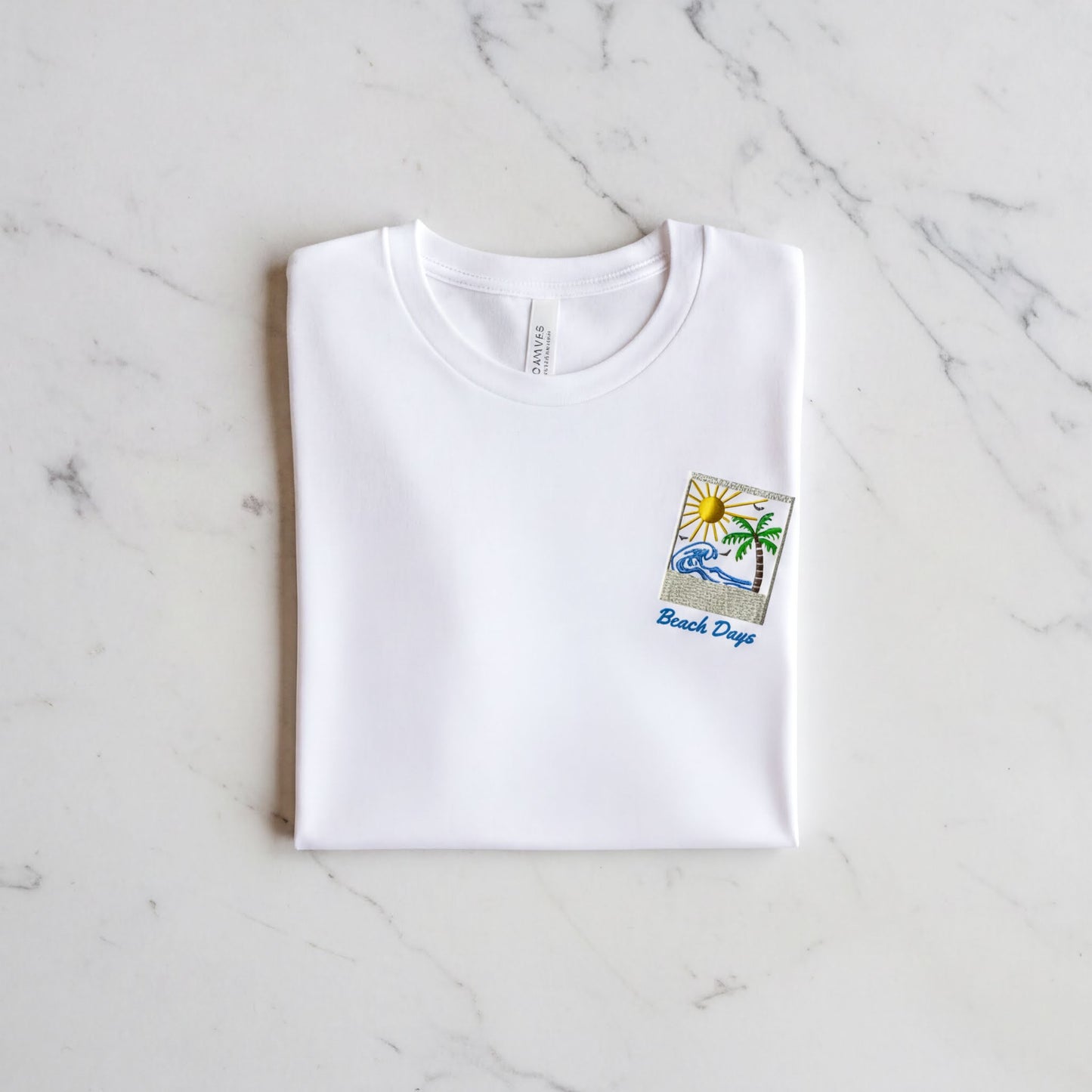 A white t-shirt with a beach-themed design and the text "Beach Days" printed on it, laid out on a marble surface.
