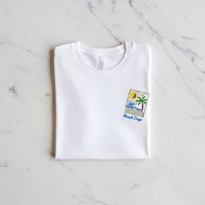 A white t-shirt with a beach-themed design and the text "Beach Days" printed on it, laid out on a marble surface.