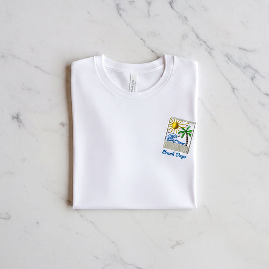 A white t-shirt with a beach-themed design and the text "Beach Days" printed on it, laid out on a marble surface.