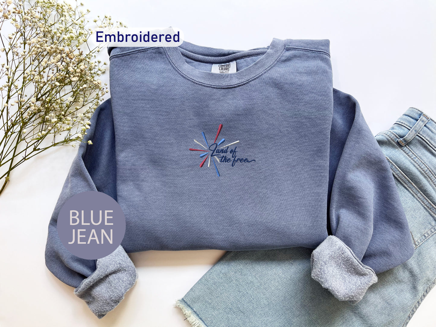 a blue sweatshirt with an embroidered design, a pair of blue jeans, and some flowers on a white background.