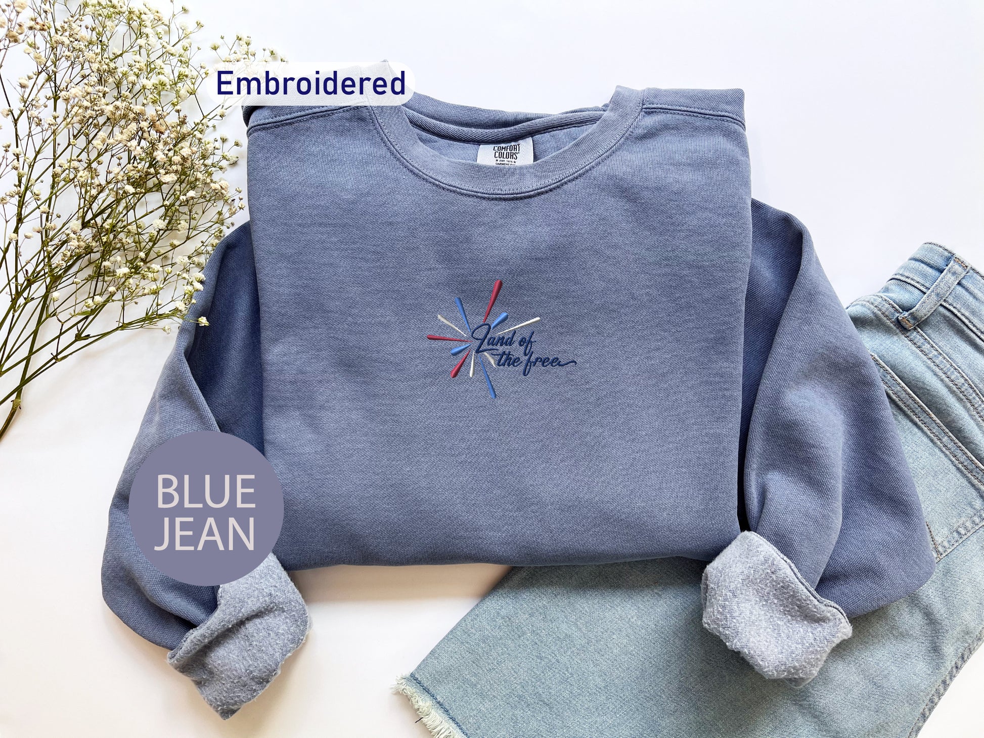 a blue sweatshirt with an embroidered design, a pair of blue jeans, and some flowers on a white background.