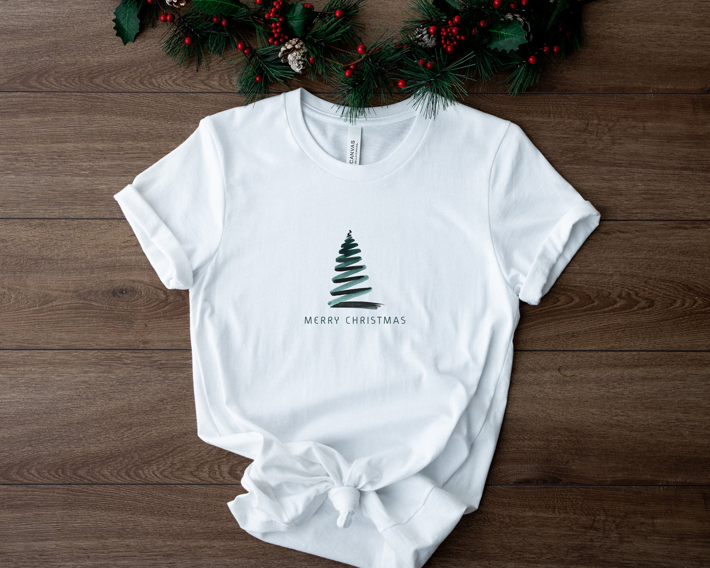 A white t-shirt with a Christmas tree design and the text "MERRY CHRISTMAS" printed on it, placed on a wooden surface with a wreath of pine branches and red berries in the background.