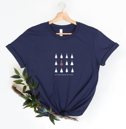 A navy blue t-shirt with a graphic design of white Christmas trees and the text "Let your heart be light" printed on it, hanging on a wooden hanger.