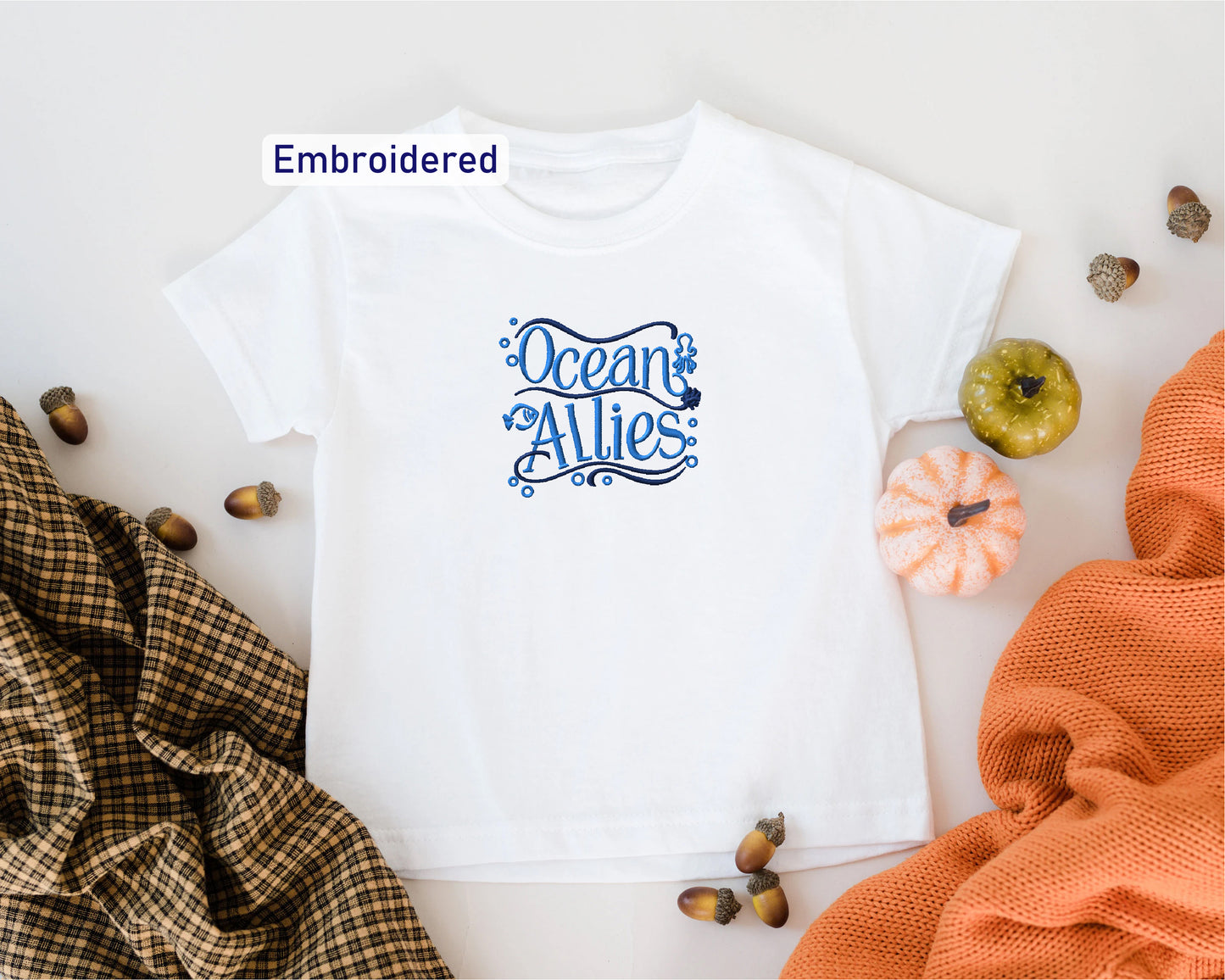 a white t-shirt with a blue embroidered design, surrounded by autumn-themed items such as acorns, pumpkins, and a plaid blanket.