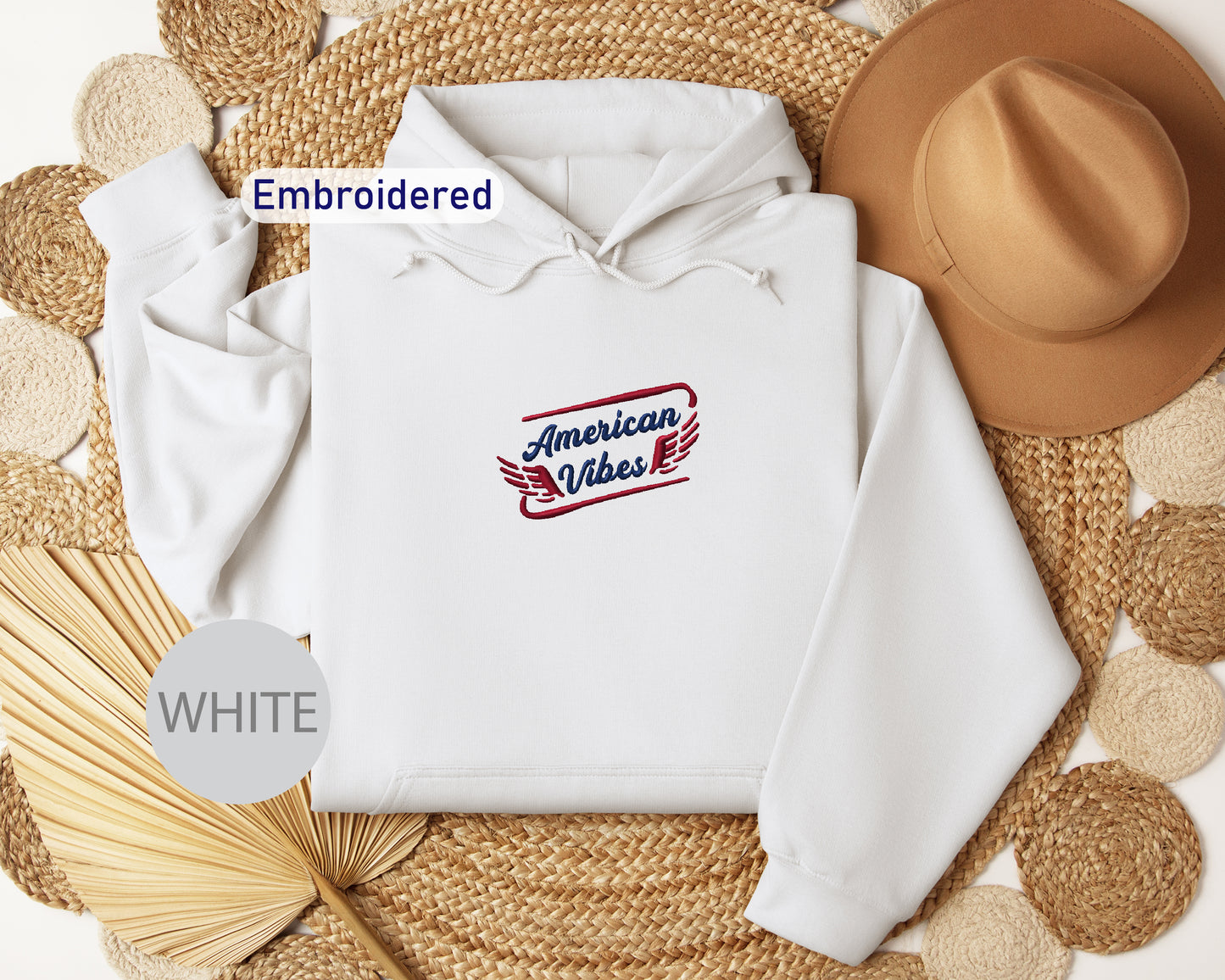 a white hoodie with an embroidered design, a brown hat, and a straw hat, all arranged on a woven mat.