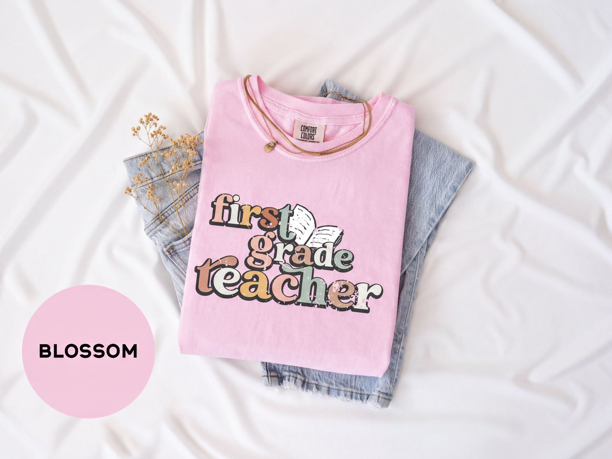 A pink t-shirt with the text "first grade teacher" printed on it, along with a necklace, is laid out on a white surface.
