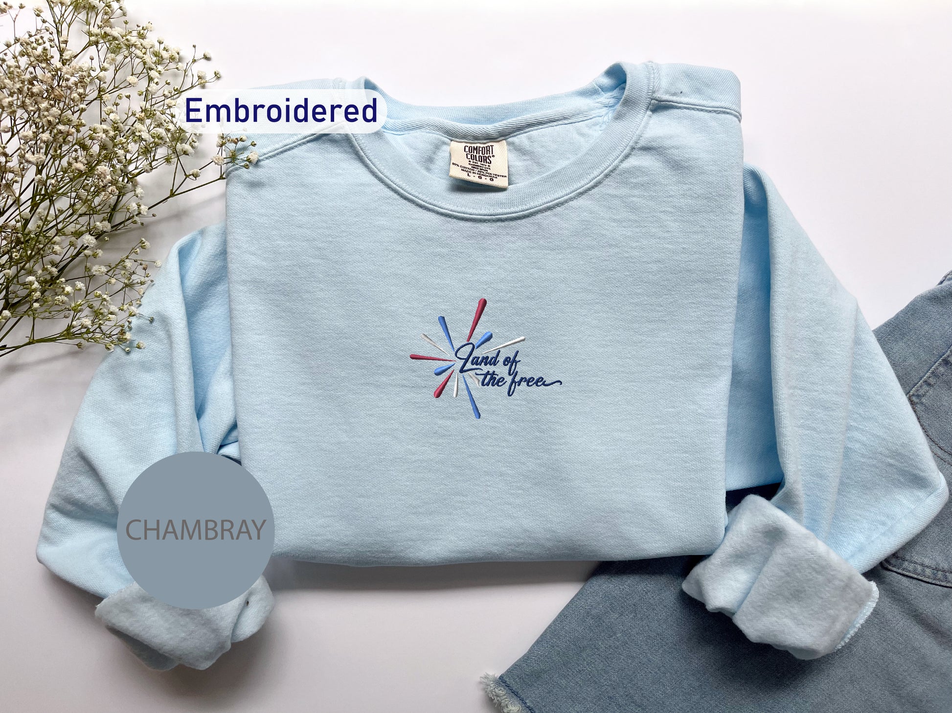 a light blue long-sleeved shirt with a patriotic design, a pair of denim jeans, and a small floral arrangement on a white background.