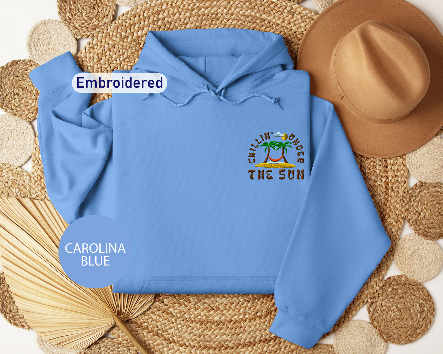 a blue hoodie with a tropical beach design, a brown hat, and a straw hat, all arranged on a woven mat.