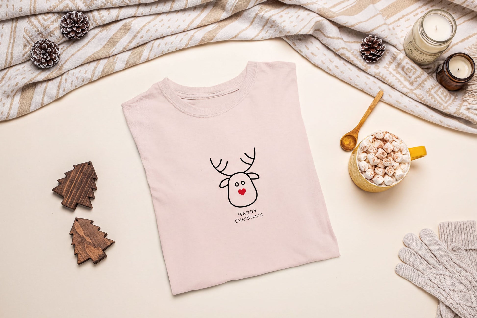 A pink t-shirt with a cartoon reindeer and the text "Merry Christmas" is displayed on a table, surrounded by holiday decorations such as pine cones, a candle, and a mug of hot chocolate.