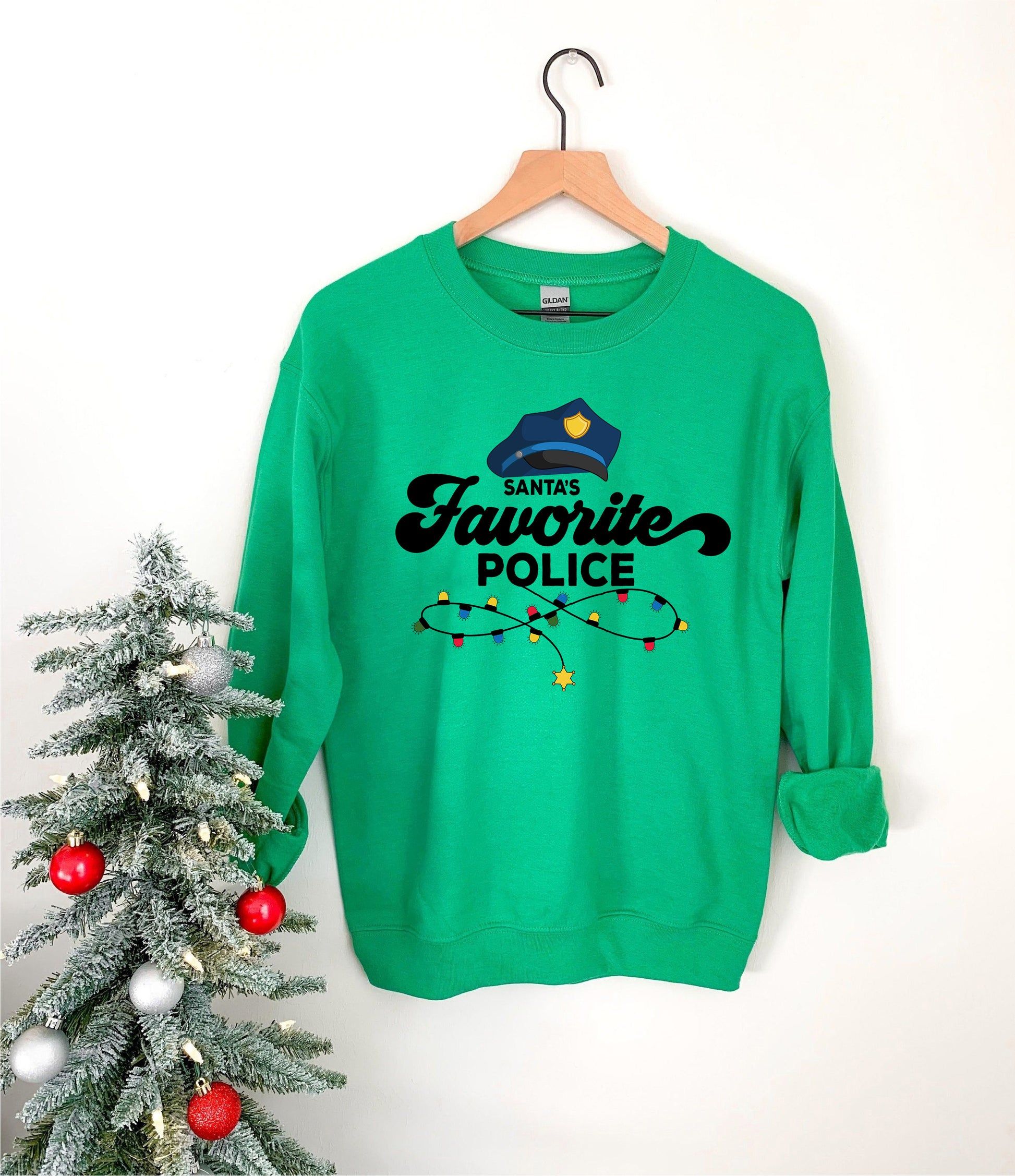 A green sweatshirt with a Santa hat and the text "Santa's Favorite Police" printed on it, hanging on a wooden hanger next to a Christmas tree decorated with ornaments.