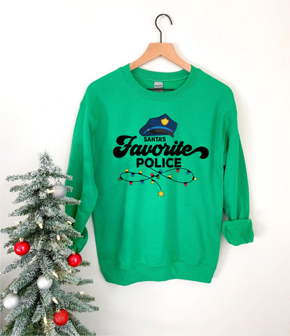 A green sweatshirt with a Santa hat and the text "Santa's Favorite Police" printed on it, hanging on a wooden hanger next to a Christmas tree decorated with ornaments.