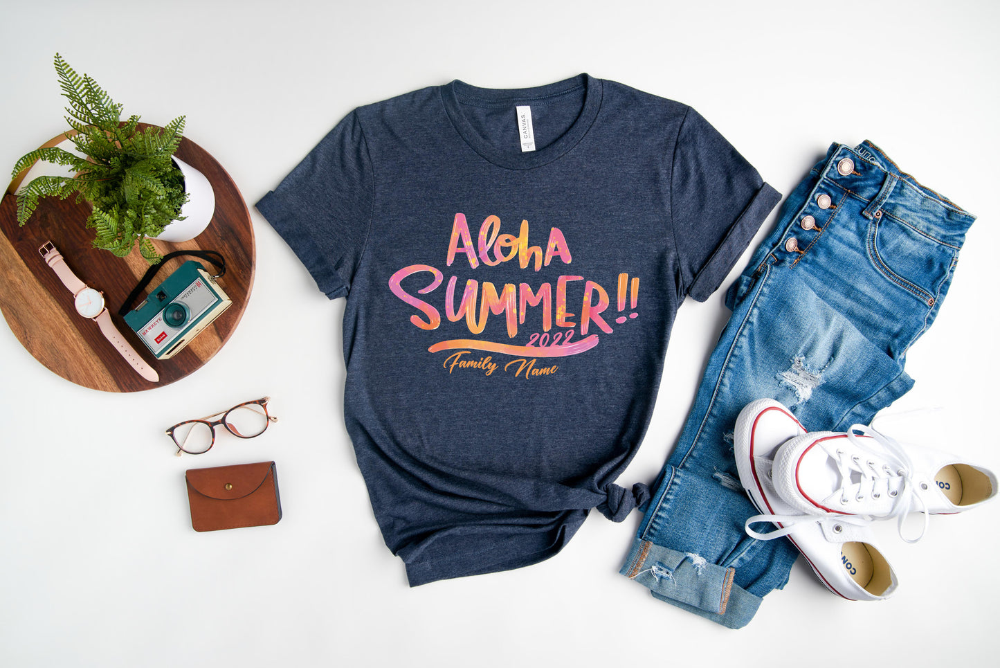 A casual summer outfit consisting of a blue t-shirt with a graphic design, jeans, and white sneakers, along with a pair of glasses, a wallet, and a potted plant, all laid out on a white background.