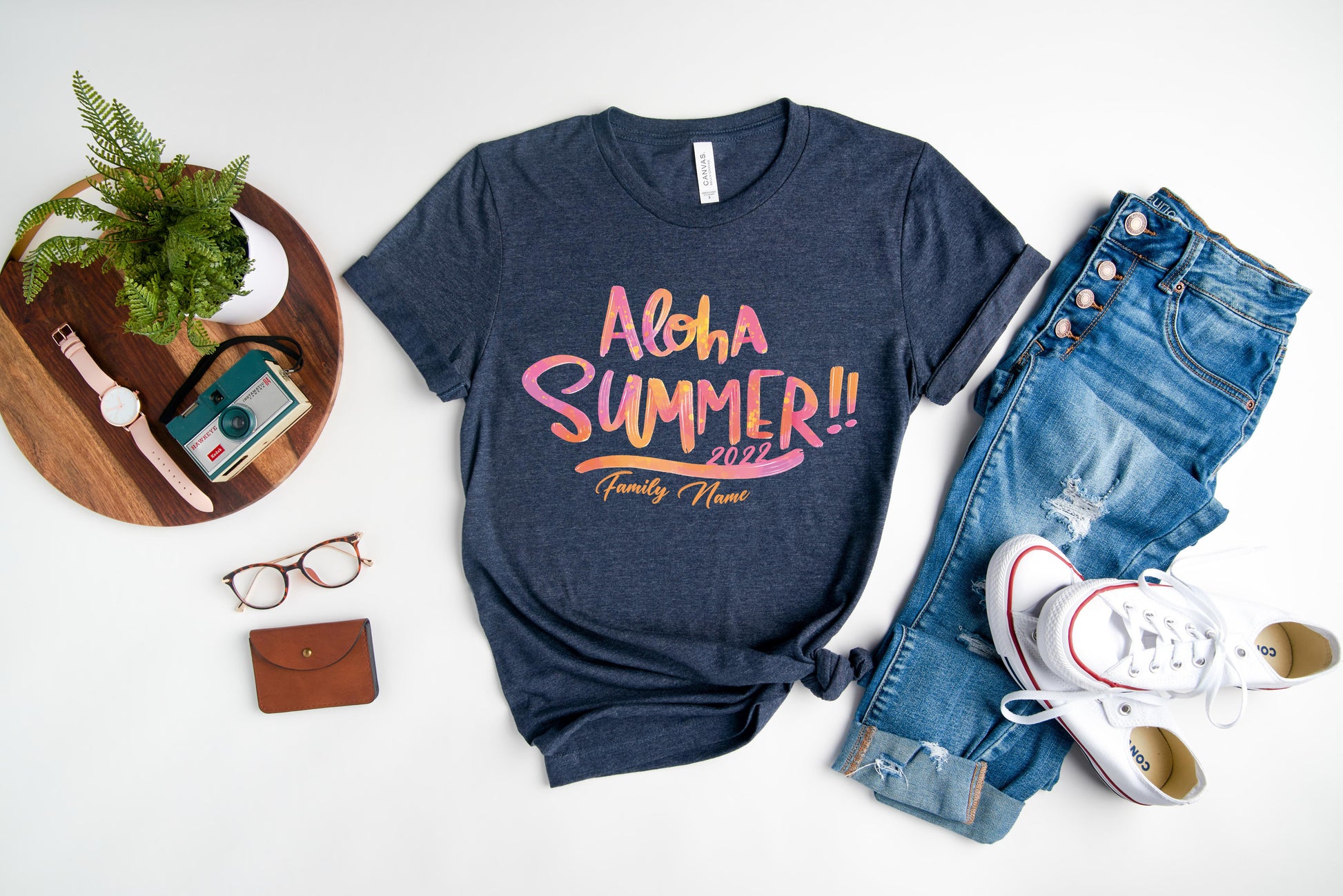 A casual summer outfit consisting of a blue t-shirt with a graphic design, jeans, and white sneakers, along with a pair of glasses, a wallet, and a potted plant, all laid out on a white background.