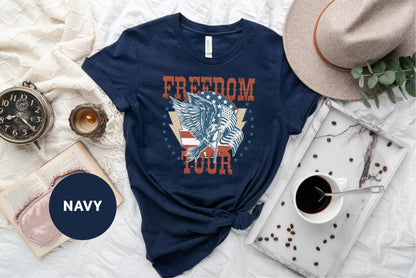 a navy blue t-shirt with a graphic design of a winged eagle and the word "FREEDOM TOUR" printed on it, along with a hat, a book, a cup of coffee, and some coffee beans.