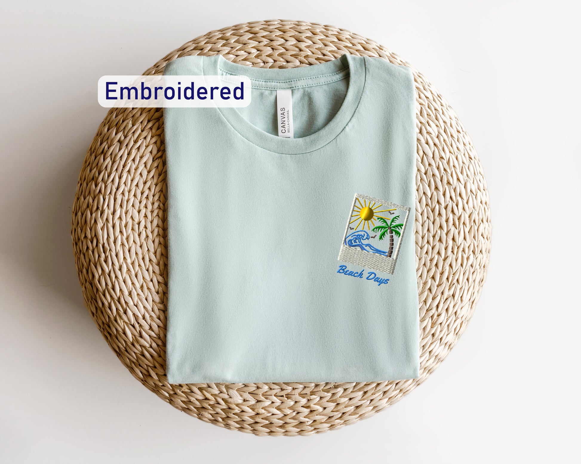 A light blue t-shirt with a beach-themed embroidered patch is displayed on a woven basket.