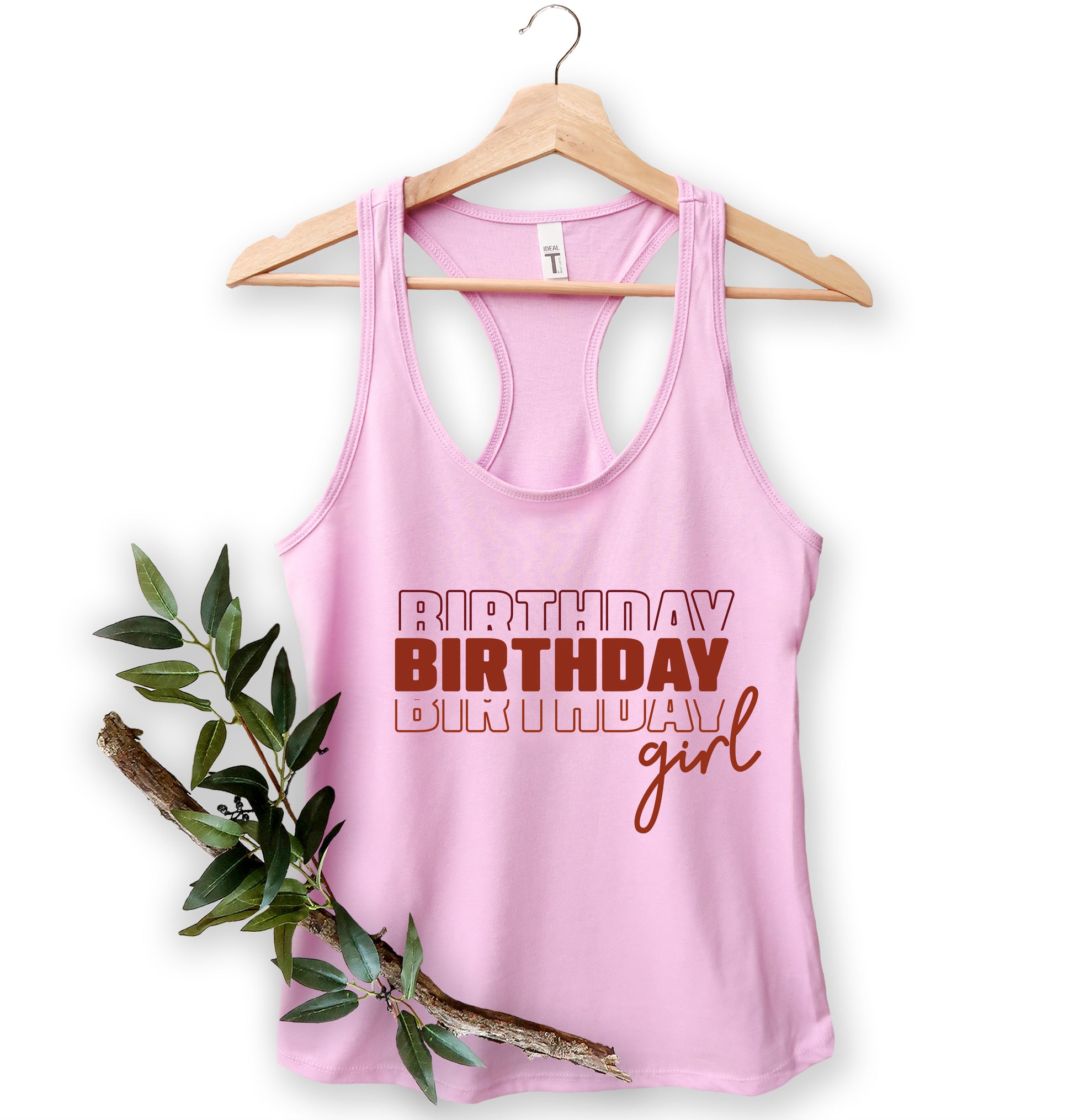 A pink tank top with the text "BIRTHDAY BIRTHDAY GIRL" printed on it, hanging on a wooden hanger.
