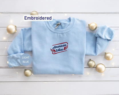a light blue long-sleeved shirt with a red and blue embroidered logo, placed on a white wooden surface alongside gold ornaments and a string of lights.