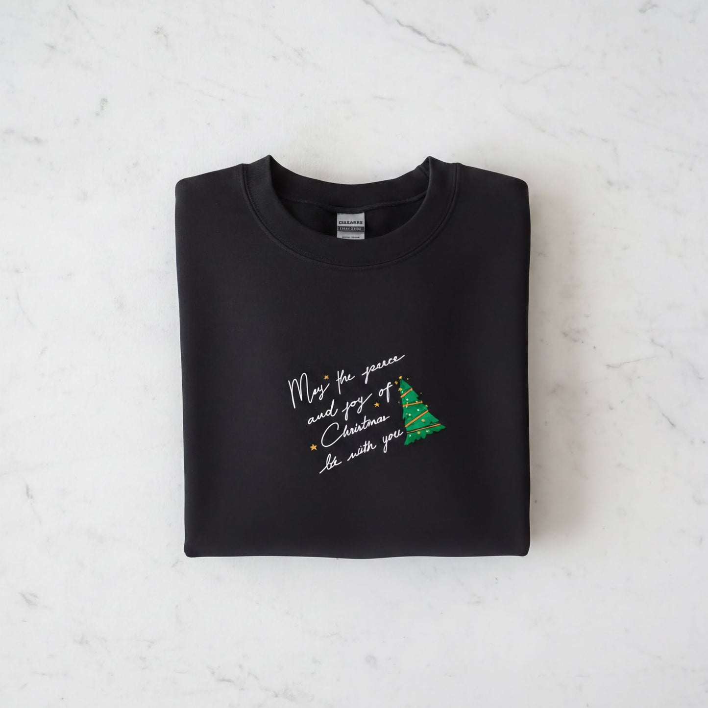 A black sweatshirt with a Christmas tree design and a message printed on it, laid out on a white marble surface.