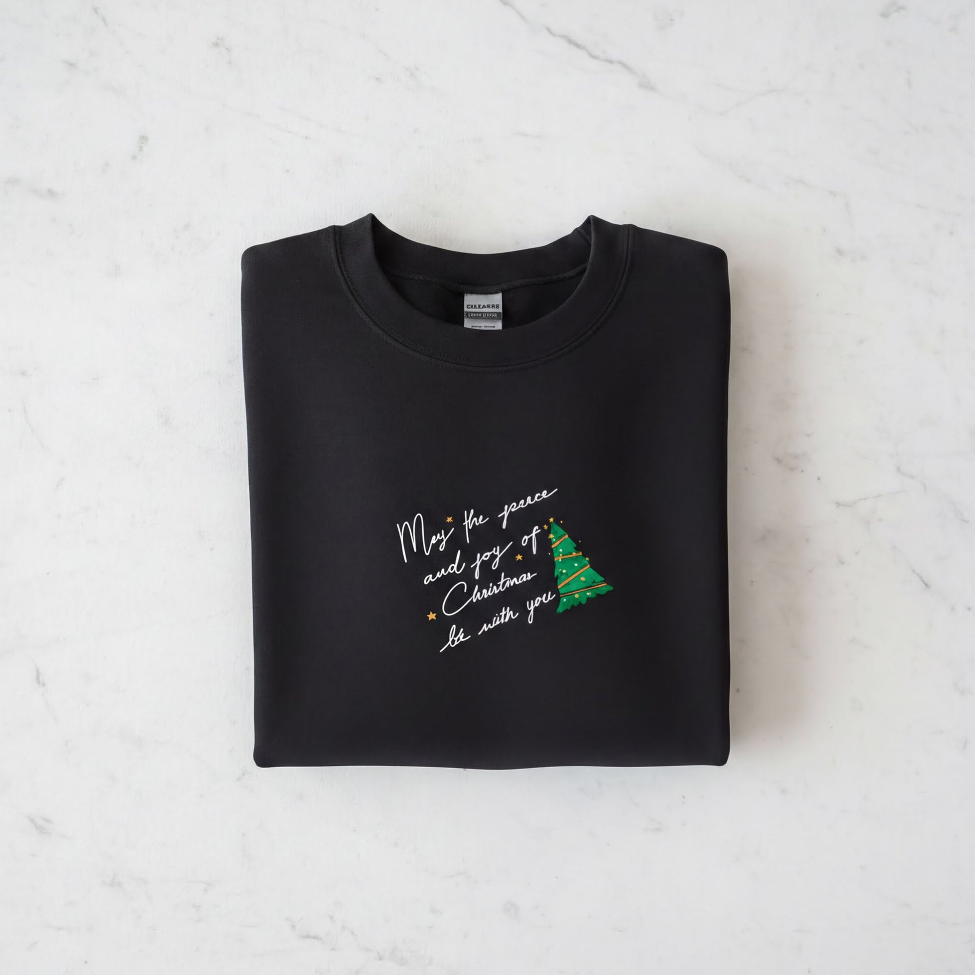 A black sweatshirt with a Christmas tree design and a message printed on it, laid out on a white marble surface.