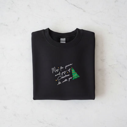 A black sweatshirt with a Christmas tree design and a message printed on it, laid out on a white marble surface.