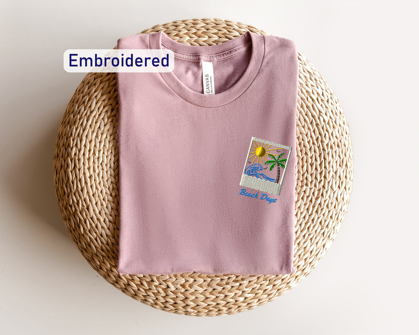 a pink t-shirt with a beach-themed embroidered design, including a palm tree and a sun, placed on a woven basket or round pillow.