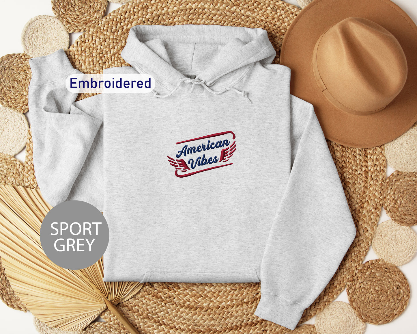 a grey hoodie with an embroidered logo, a brown hat, and straw hats arranged on a woven mat.
