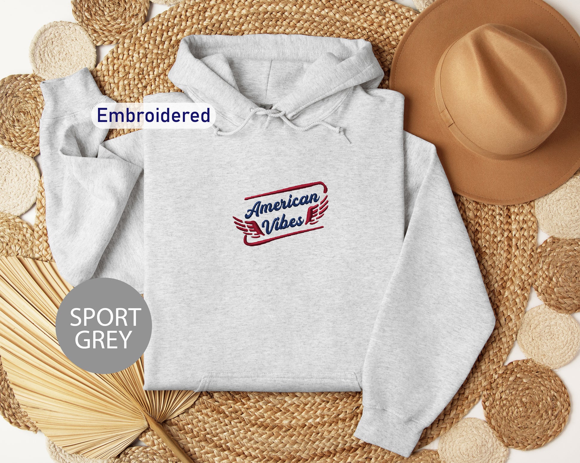 a grey hoodie with an embroidered logo, a brown hat, and straw hats arranged on a woven mat.