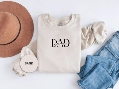 A casual outfit consisting of a white sweatshirt with the text "DAD" printed on it, a brown hat, and a pair of blue jeans is laid out on a white background.