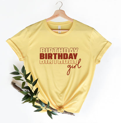 A yellow t-shirt with the text "BIRTHDAY BIRTHDAY girl" printed on it, along with a wooden hanger.