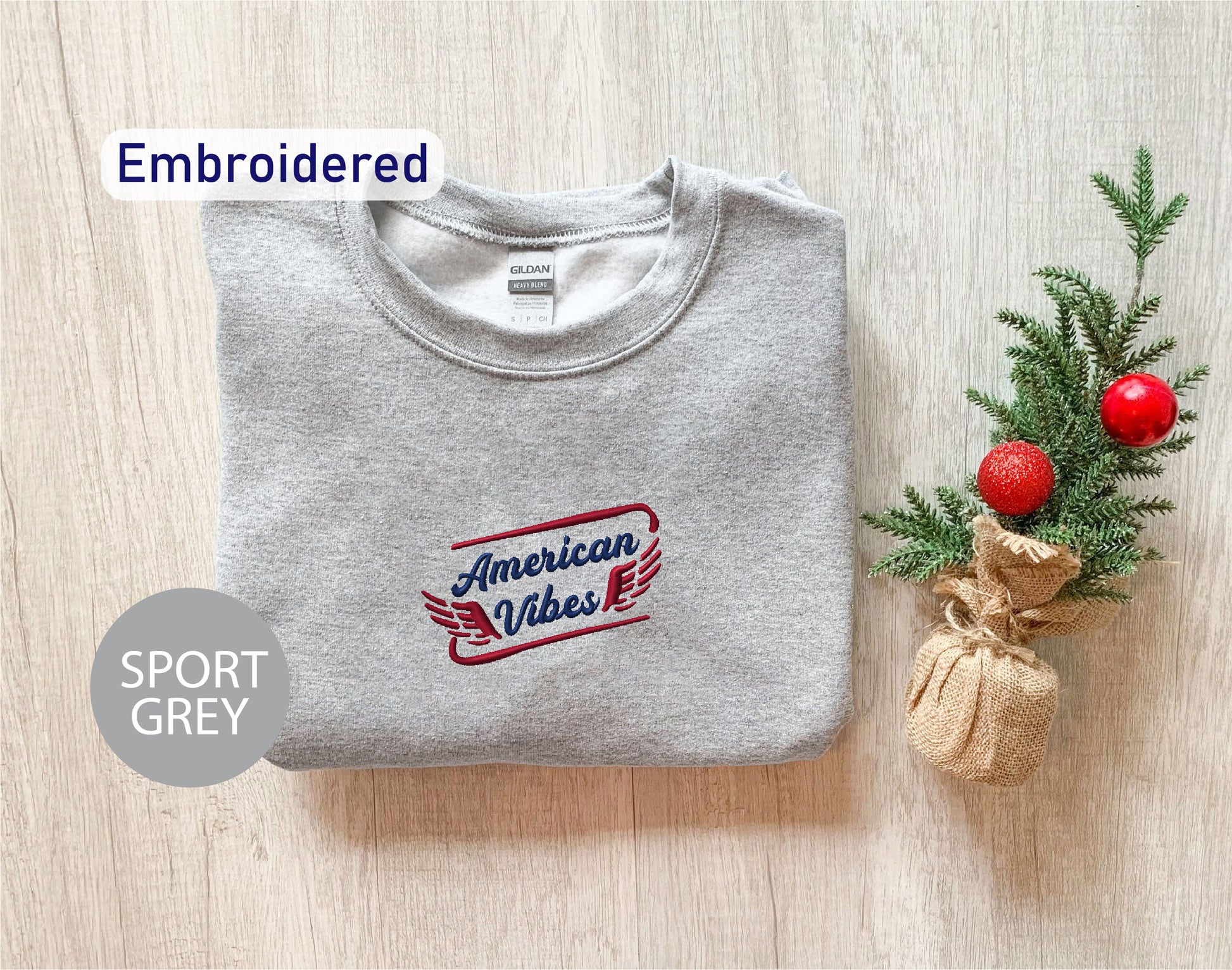 a grey sports sweatshirt with the text "American Vibes" embroidered on it, along with a small Christmas tree and a burlap sack.