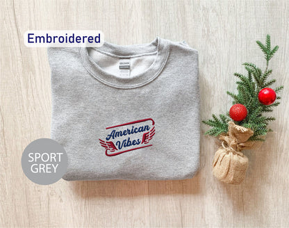 a grey sports sweatshirt with the text "American Vibes" embroidered on it, along with a small Christmas tree and a burlap sack.