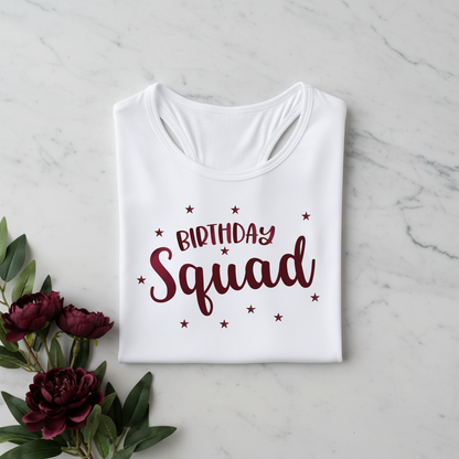 A white t-shirt with the text "Birthday Squad" printed on it, along with some stars, is displayed on a marble surface next to a bouquet of red flowers.