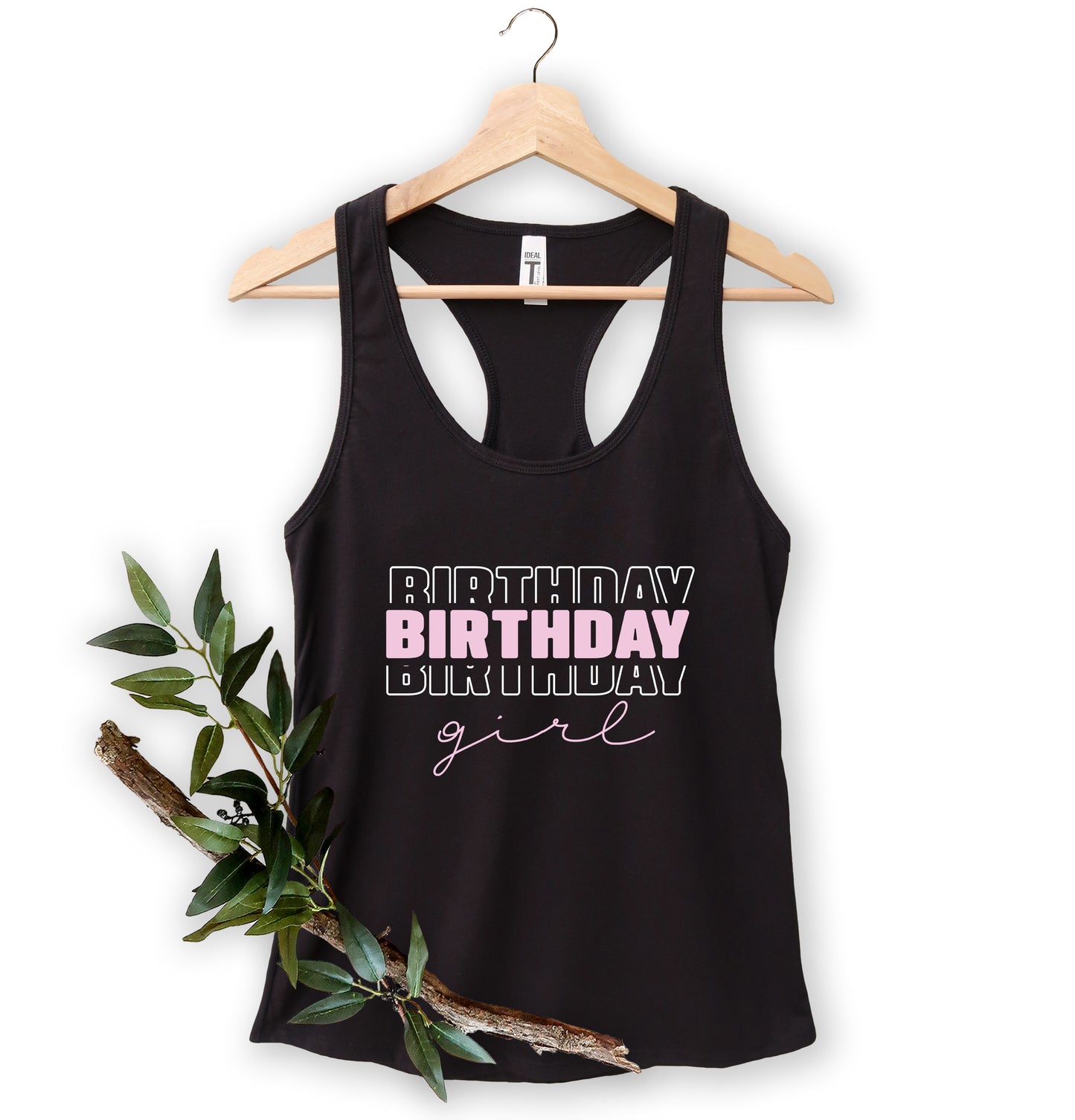 A black tank top with the text "BIRTHDAY GIRL" printed on it, hanging on a wooden hanger.