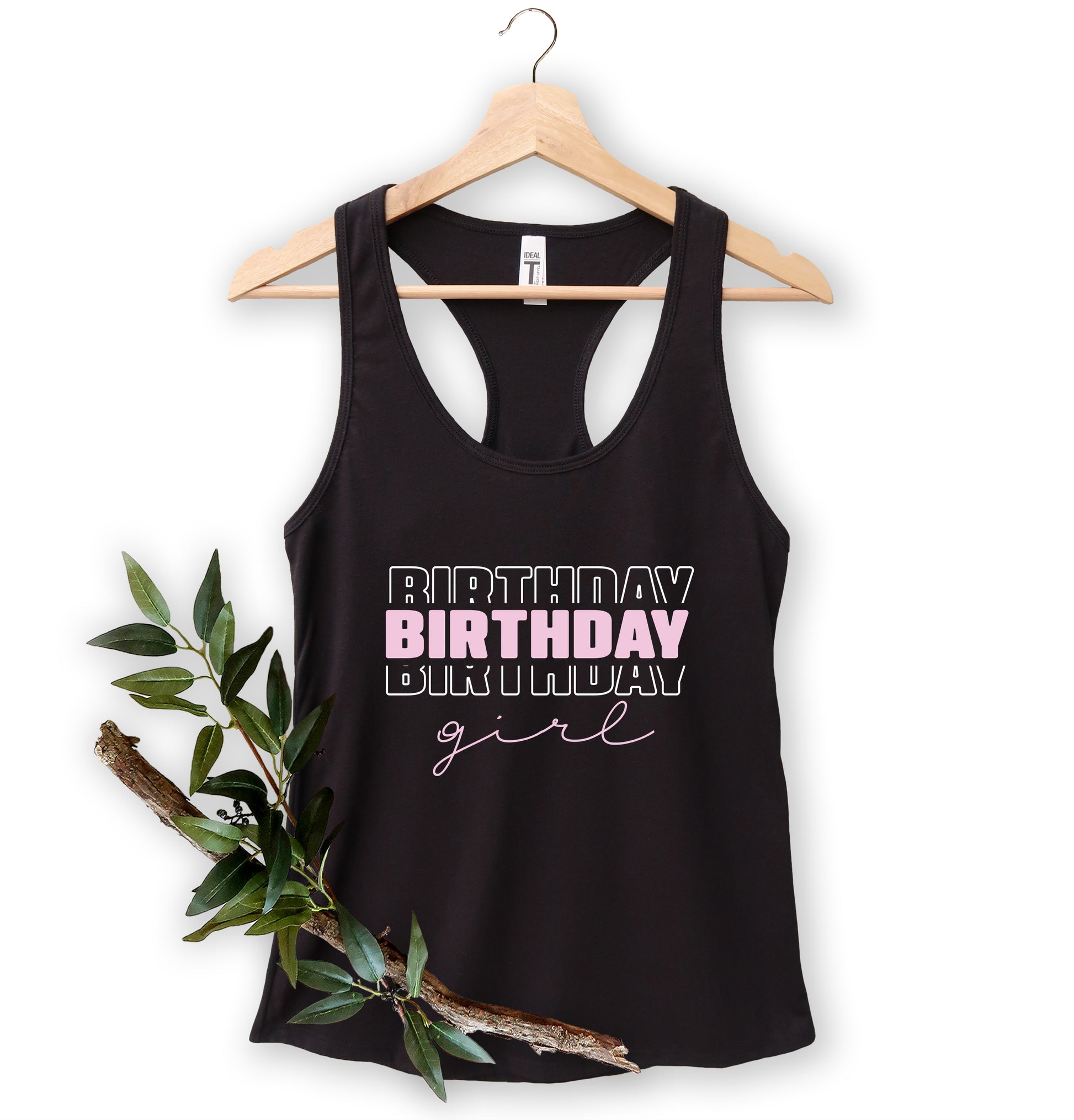 A black tank top with the text "BIRTHDAY GIRL" printed on it, hanging on a wooden hanger.