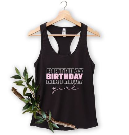A black tank top with the text "BIRTHDAY GIRL" printed on it, hanging on a wooden hanger.