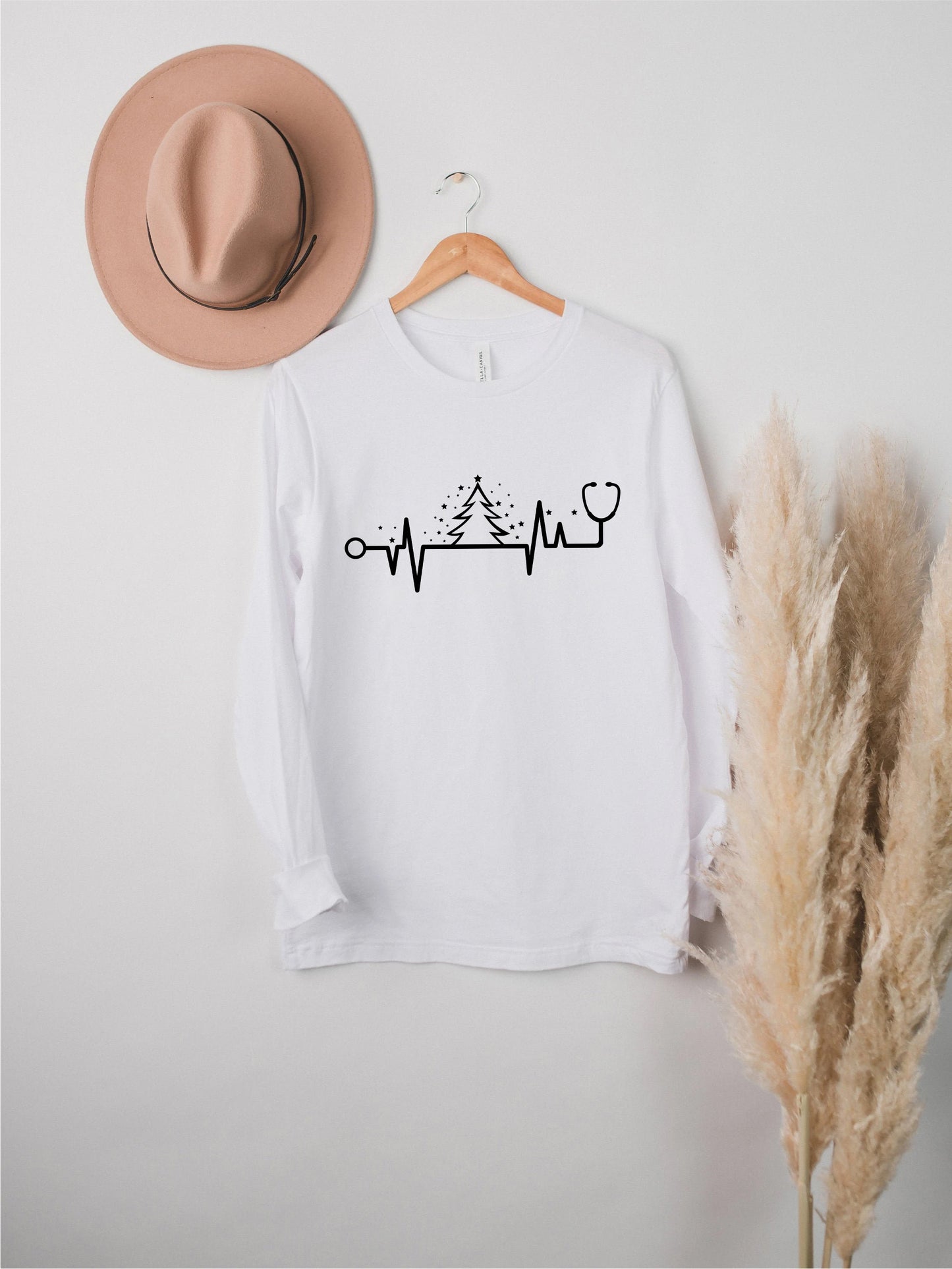 A white long-sleeved t-shirt with a graphic design of a stethoscope and a line graph, along with a pink hat and potted plant, all displayed on a white wall.