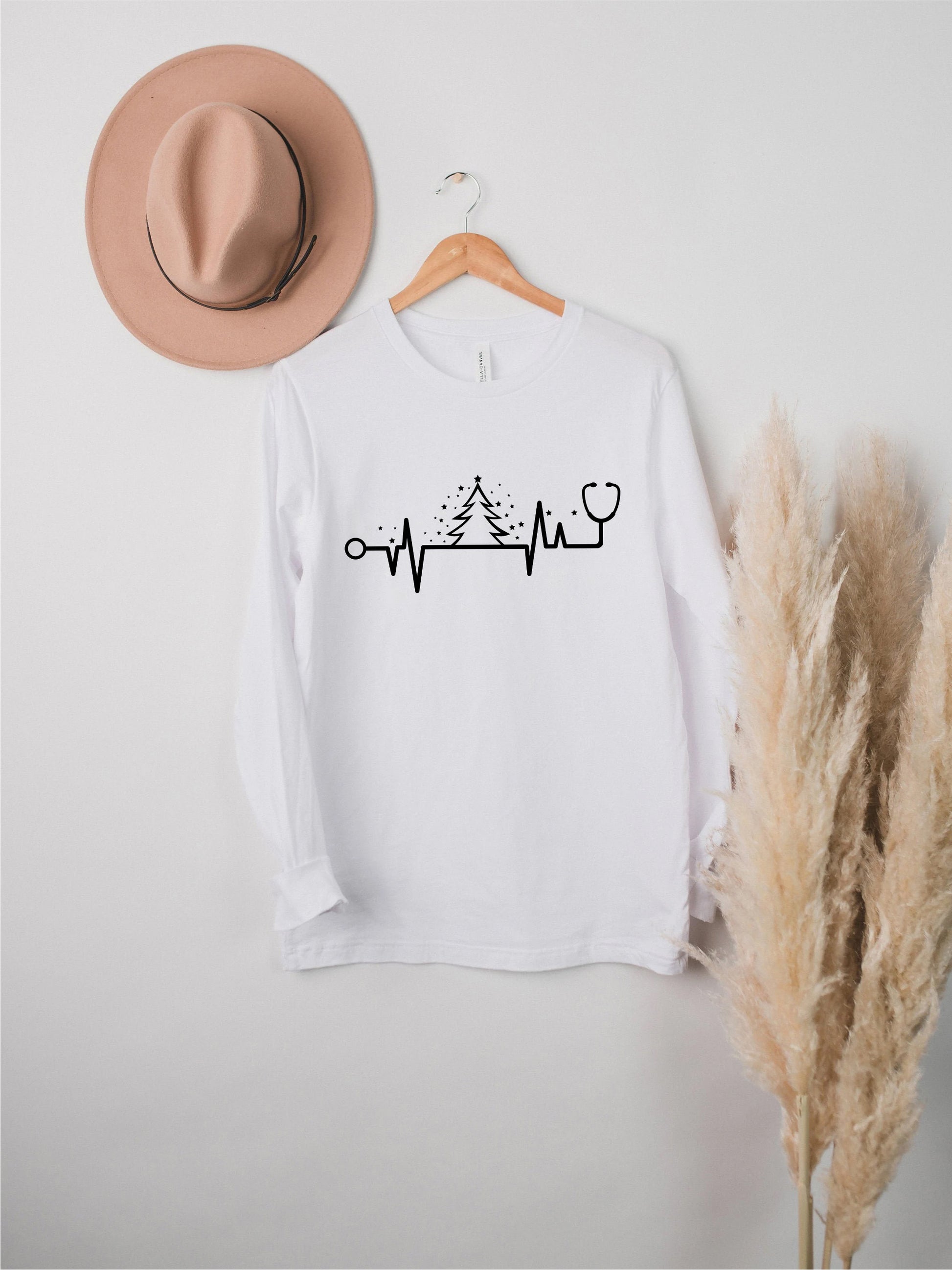 A white long-sleeved t-shirt with a graphic design of a stethoscope and a line graph, along with a pink hat and potted plant, all displayed on a white wall.