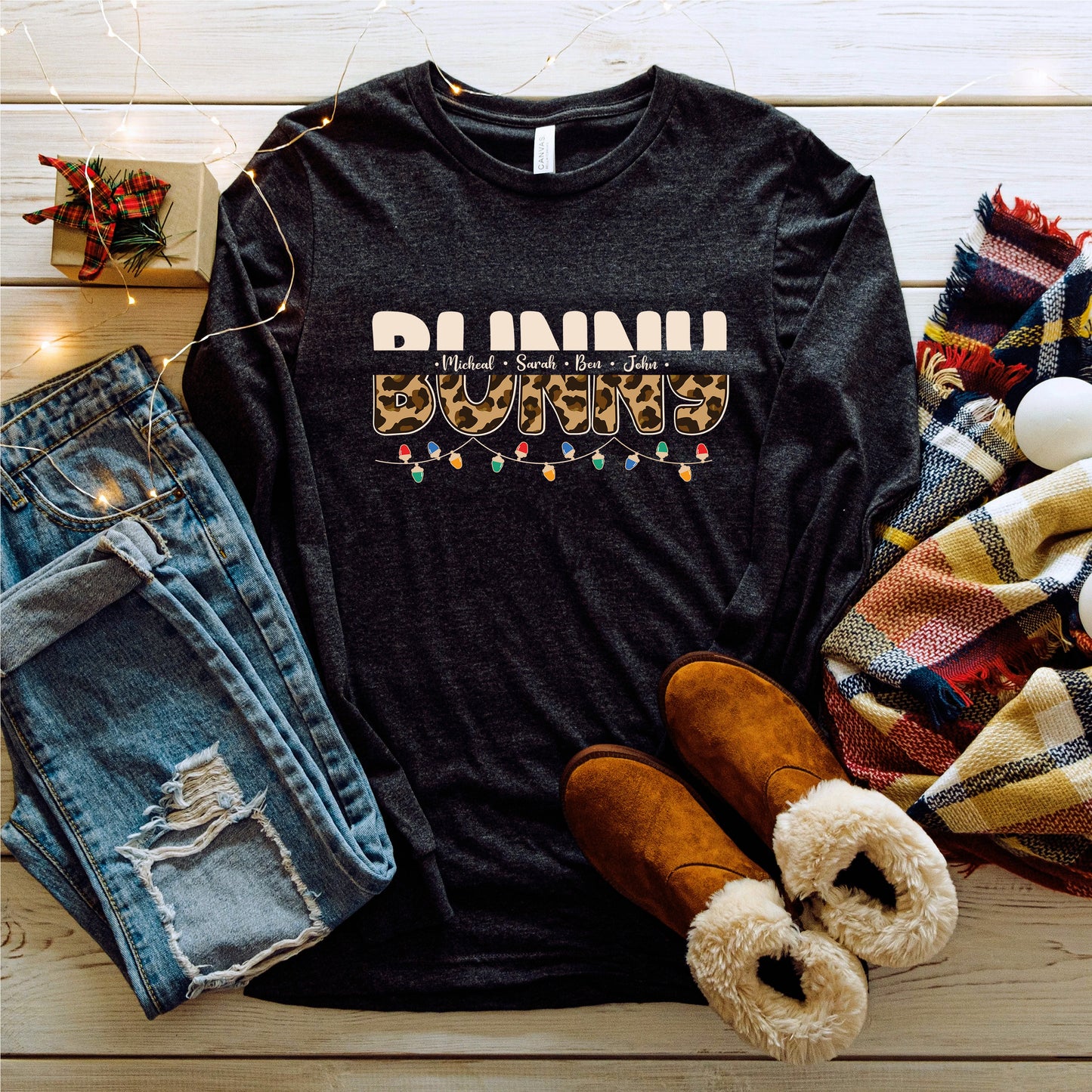 A casual outfit consisting of a black long-sleeved shirt with a leopard print design, distressed blue jeans, brown boots, and a plaid scarf is laid out on a wooden surface, along with some string lights and a small gift box.