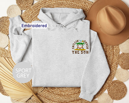 a grey hoodie with a tropical beach design, a brown hat, and a straw hat, all arranged on a woven mat.