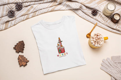 A white t-shirt with a Christmas-themed graphic, a mug of hot chocolate, pine cones, and a pair of gloves are arranged on a beige background.