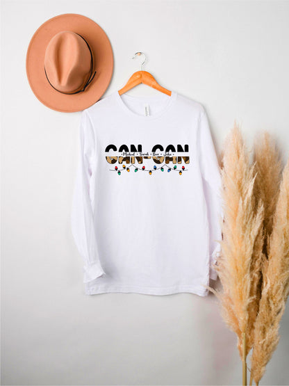 A white long-sleeved t-shirt with the text "CAN CAN" printed on it, hanging on a wooden hanger. The t-shirt is displayed against a white wall, with a brown hat and some potted plants visible in the background.