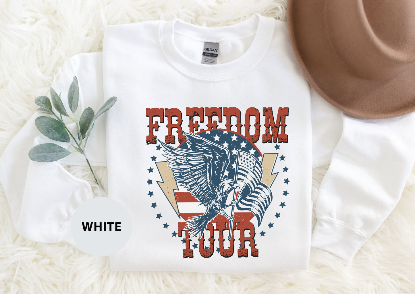 A white long-sleeved t-shirt with a graphic design featuring a bird, an eagle, and an American flag, along with the text "FREEDOM TOUR" printed on it. The t-shirt is laid out on a white surface, and there is a brown hat placed next to it.